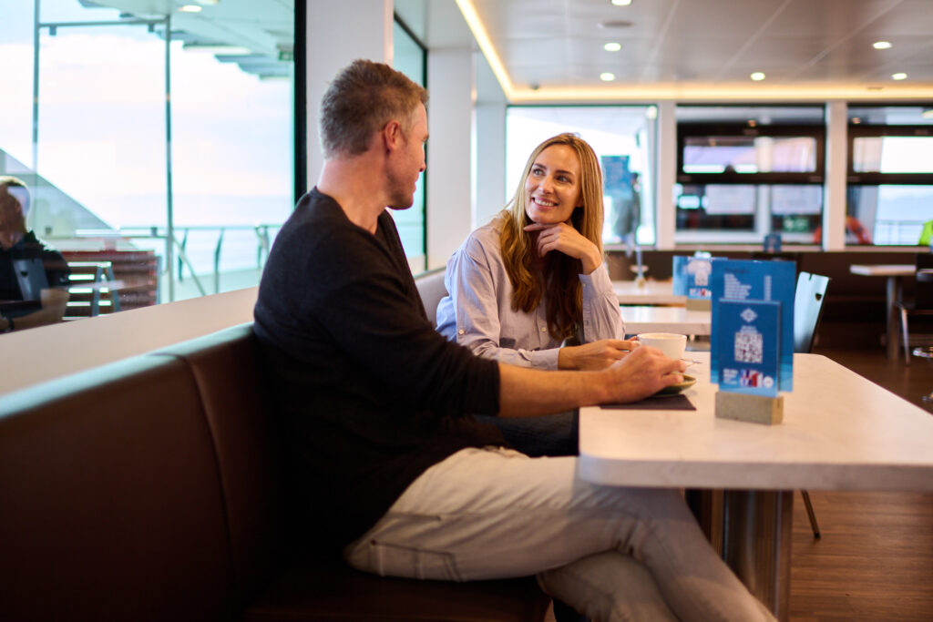 Zwei Personen sitzen an einem Tisch in einem modernen Innenraum, möglicherweise einem Café oder Restaurant. Im Hintergrund sind große Fenster mit Ausblick sowie Regale mit verschiedenen Gegenständen zu sehen.