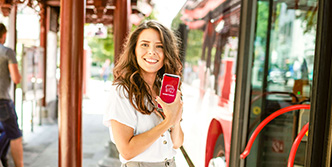 Porträt einer Frau vor dem Bus an einer Bushaltestelle. Sie schaut freundlich in die Kamera und zeigt auf ihrem Handy die Stadtwerke Konstanz App in die Kamera.