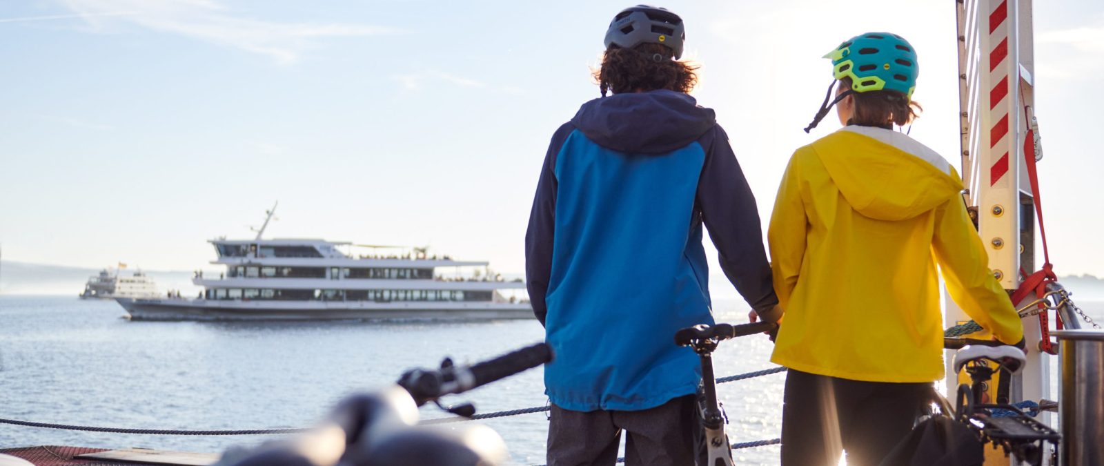 Zwei Personen stehen auf dem Fahrbahndeck eines Fährschiffs. Beide Tragen einen Fahrradhelm und stehen mit Blick auf ein vorbeifahrendes Motorschiff der BSB neben einem Fahrrad.