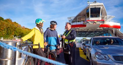 Zwei Personen mit Fahrrädern unterhalten sich mit einem Kassiermatrosen auf einem Fährschiff.